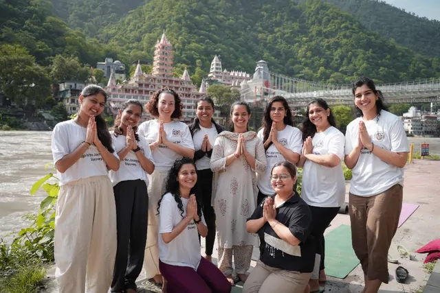 Prenatal yoga teacher training group in namaste by the Ganga, with Tera Manzil temple and Laxman Jhula behind them